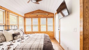 Un dormitorio con cama, paredes de madera, ventilador de techo y ventanas grandes.