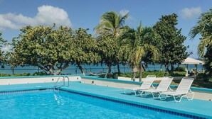 A swimming pool with clear blue water, surrounded by white lounge chairs and palm trees. - Beachfront Sunset Oasis  Adult-Only, Pool & Views (Frederiksted)