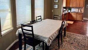 Un coin repas avec une table dressée pour quatre personnes, encadré par deux fenêtres équipées de stores, une cuisine avec des meubles en bois et un tapis à motifs sur le sol.