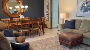Uma sala de estar com um sofá marrom, um tapete estampado, uma mesa de jantar de madeira com cadeiras e um candelabro.