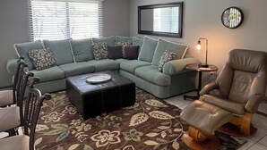 Uma sala de estar com sofá seccional, mesa de centro, poltrona de couro e tapete estampado.