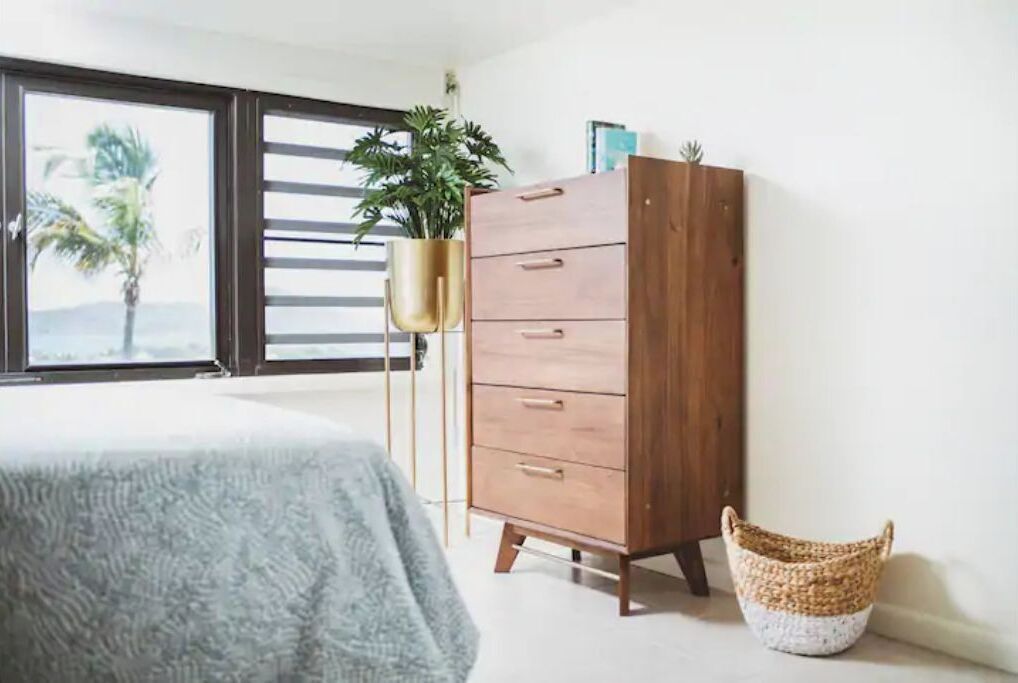 A bedroom with a bed, a wooden dresser, a hanging plant, and a woven basket.
