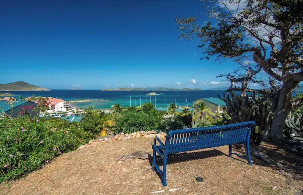 A bench overlooking a coastal view with a clear sky, a distant island, and a swimming pool area.