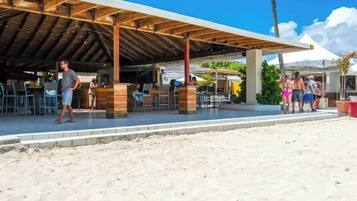 An outdoor seating area with wooden tables and chairs, a covered patio, and a sandy beach in the background.
