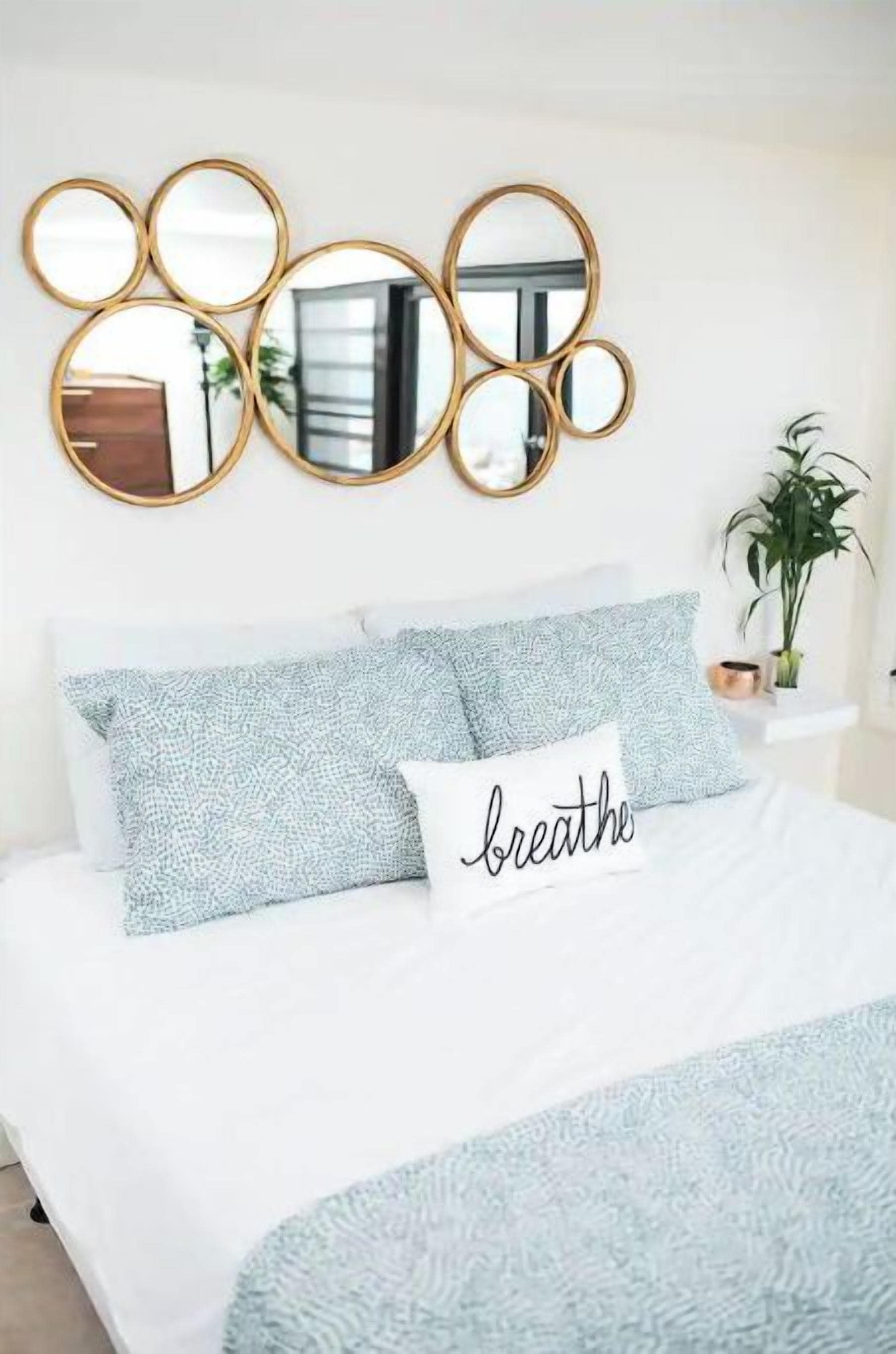 Una cama bien hecha con una colcha blanca y azul claro estampada, un cojín con la palabra 'breathe' y un conjunto de espejos montados en la pared.