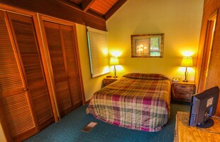 A bedroom with a bed, two bedside lamps, a desk with a computer, a window with blinds, and a wooden wardrobe.