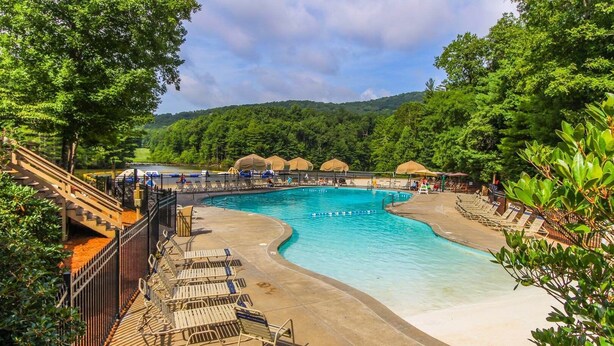 A large outdoor swimming pool with a curved design, surrounded by lounge chairs and umbrellas, set against a backdrop of lush greenery and hills. - Golf Club Villas (Marble Hill)