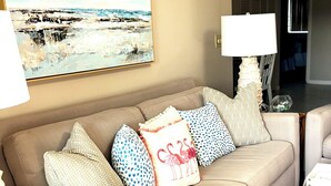 En stue med en beige sofa, mønstrede puder, et glasbord og et billede på væggen.