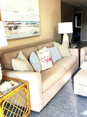 A living room with a beige sofa, patterned cushions, a glass coffee table, and a painting on the wall.