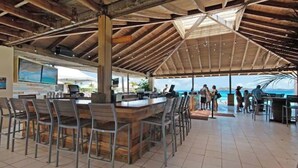 An outdoor bar area with a wooden ceiling and tiled floor, featuring a long counter with chairs and a view of the ocean.