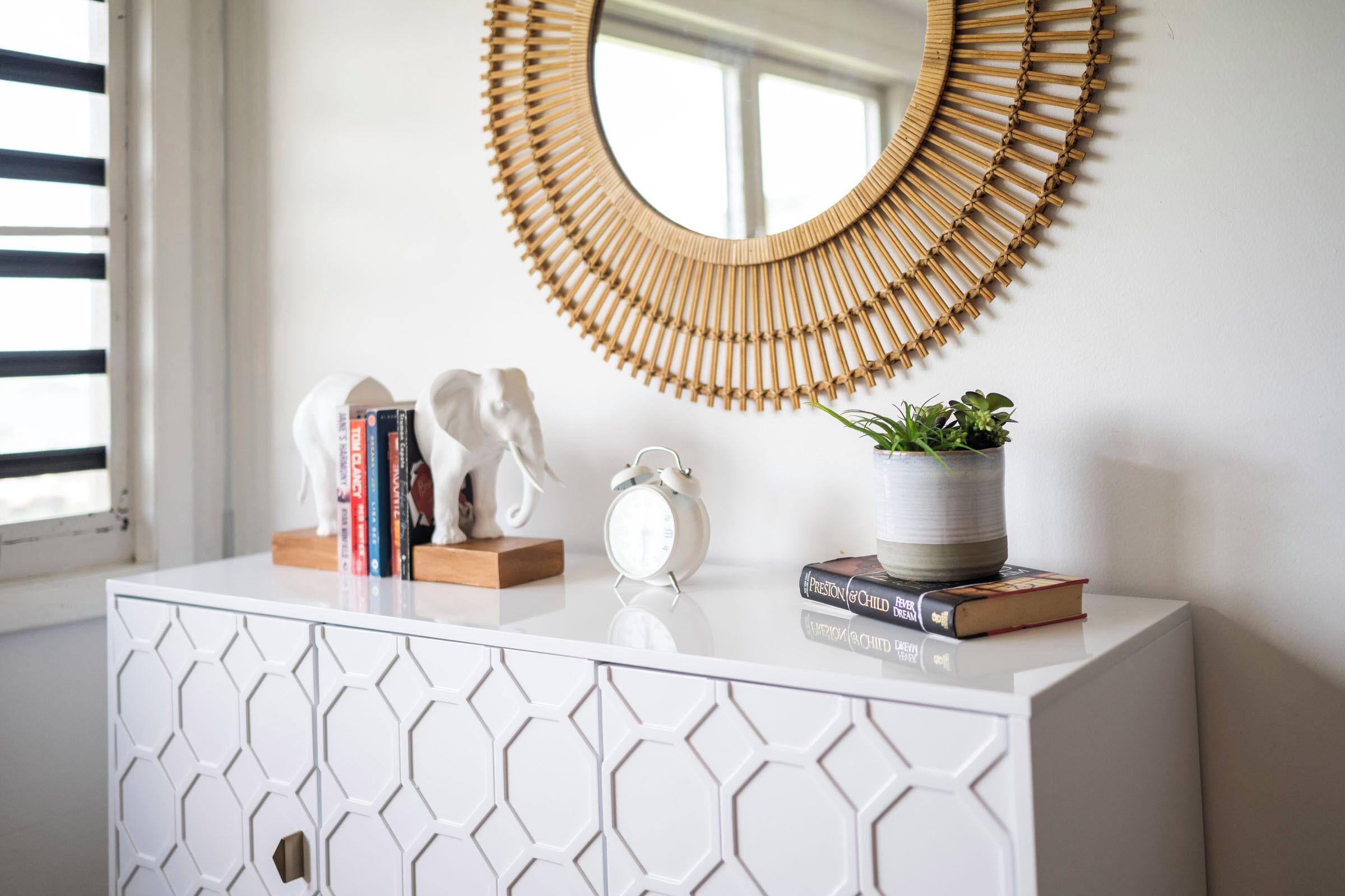 A white cabinet with a geometric pattern, a round mirror with a bamboo frame, a small potted plant, a white alarm clock, and a few books.