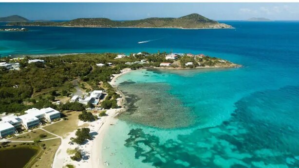 Luftaufnahme einer Küstenregion mit Strand, klarem türkisblauem Wasser und einer kleinen Insel mit Gebäuden.