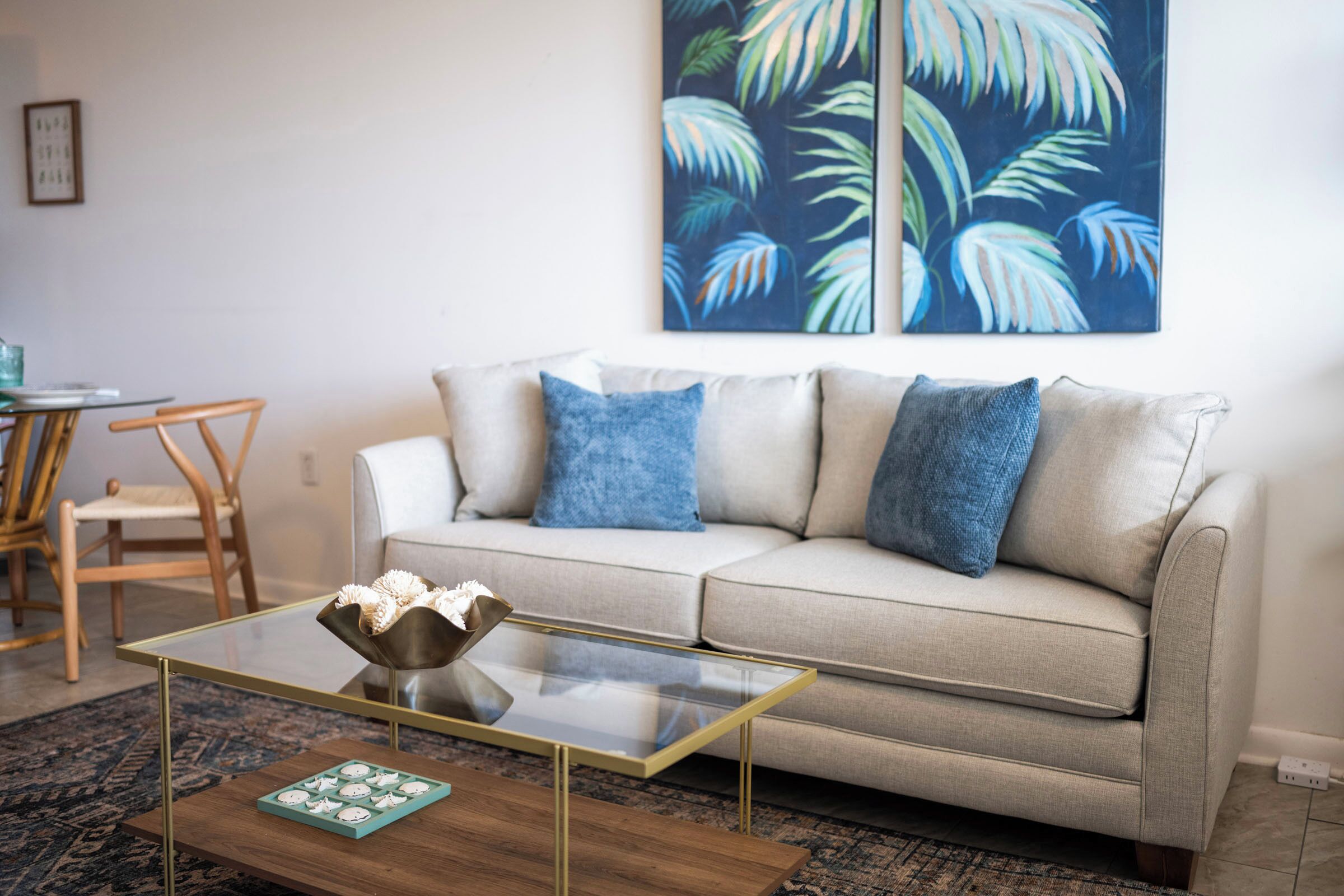 Un soggiorno con un tavolino in vetro, un divano beige con cuscini blu, una sedia in legno e un quadro con una foglia tropicale in cornice appeso al muro.