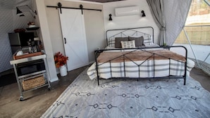 Una habitación con carpa, una cama, una mesita con una bandeja, un aire acondicionado de pared y una ventana con cortinas.
