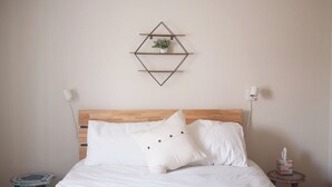 Een net opgemaakt bed met witte lakens en twee kussens, een houten hoofdbord en een geometrisch wandplankje met een plantje.