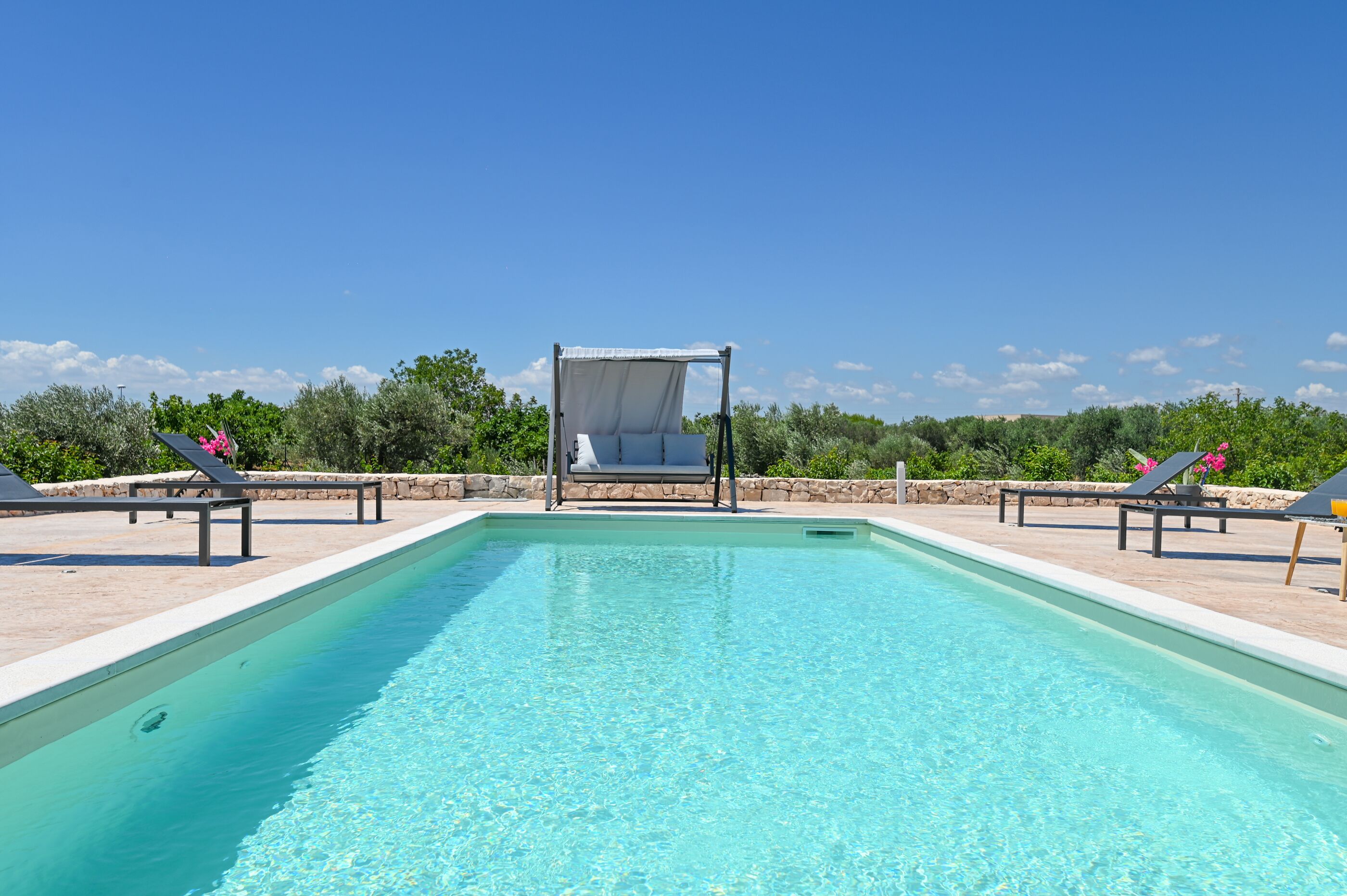 A rectangular swimming pool with clear blue water, surrounded by lounge chairs and a canopy structure.
