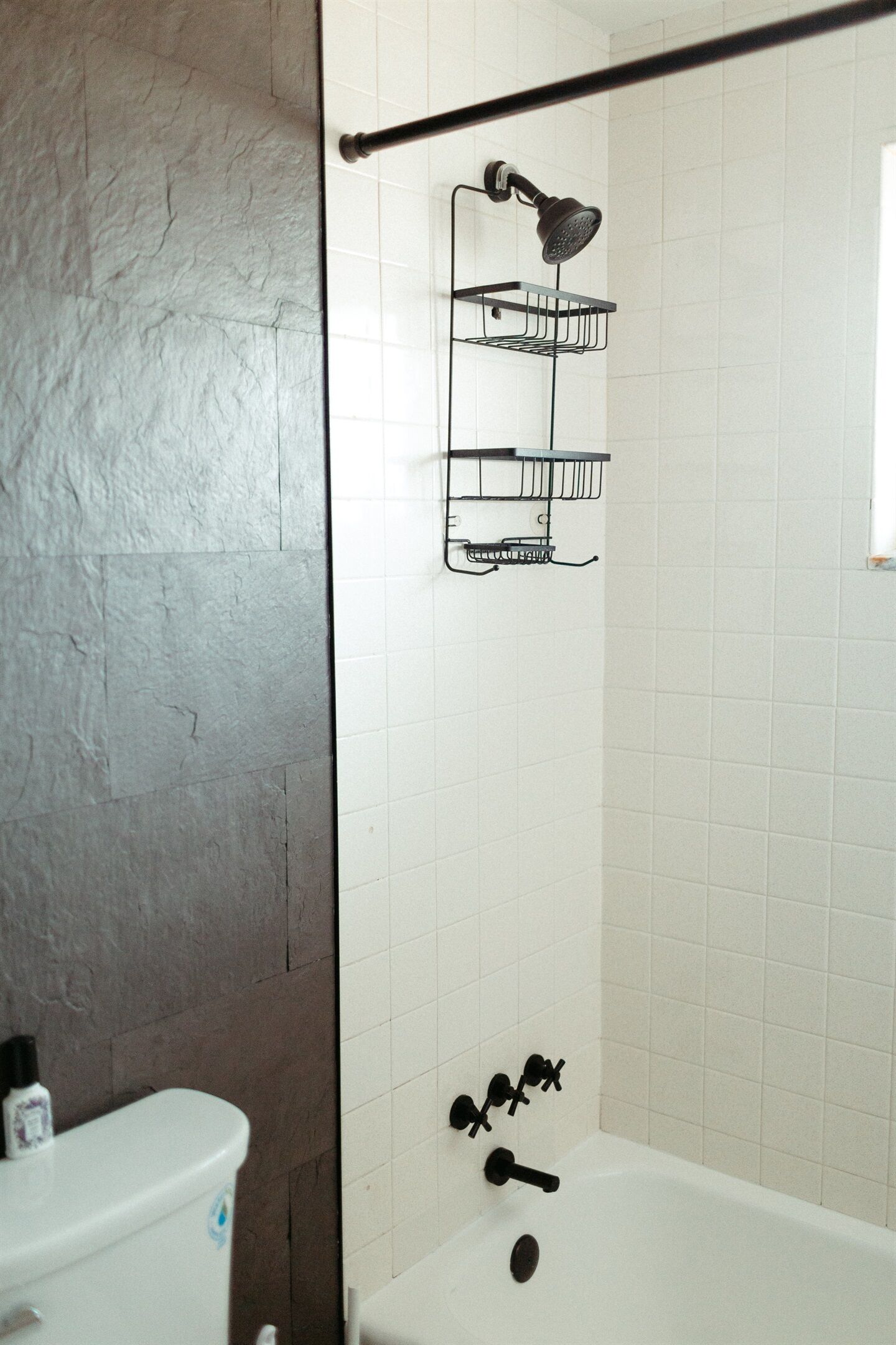 Een badkamer met een witte badkuip, een douchekop en een toilet. Er zitten zwarte haken aan de muur en een witte fles op het toilet.