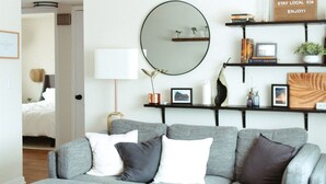 A modern living room with a grey sectional sofa, a round mirror, and a coffee table. There is a bookshelf with books and decorative items, and a welcome sign on the wall. - Modern, Warm, Chic Flat in Westerville (Westerville)