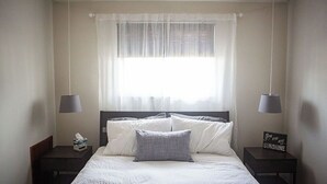 Un lit bien fait, avec une literie blanche et grise, deux tables de chevet avec des lampes, un petit panneau lumineux sur la table de chevet et une fenêtre ornée de rideaux légers.