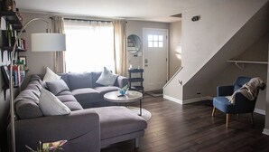 Un salon moderne avec un canapé gris, une table basse ronde et un fauteuil bleu. On y trouve une bibliothèque remplie de livres et une lampe. Un escalier est visible au fond.