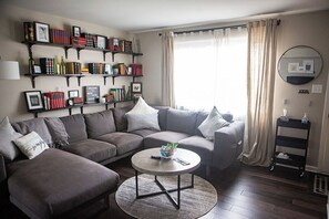 A living room with a sectional sofa, a coffee table, and a bookshelf filled with books and decorative items. - Bright, Modern Townhome with Natural Light (Westerville)