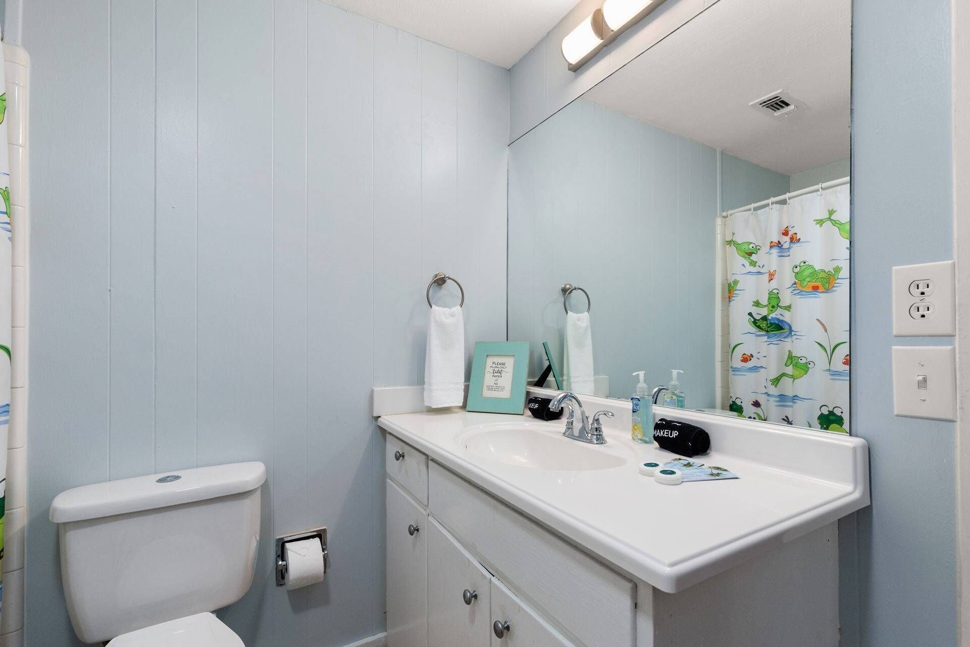 Ein Badezimmer mit Toilette, Waschbecken und Spiegel. Ein Handtuchhalter mit Schild, eine Flasche Handseife und eine schwarze Kosmetiktasche stehen am Waschbecken.