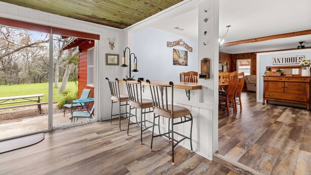 A rustic interior with a wooden ceiling, a dining area with bar stools, a kitchen with wooden cabinets, and a view of the outdoors.
