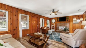 A cozy living room with wooden walls, a fireplace, a ceiling fan, and a TV mounted above it.
