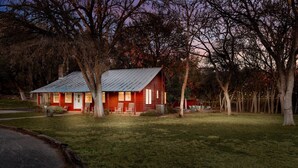 A small, single-story cabin with a metal roof, surrounded by a grassy yard and trees.