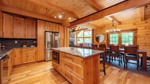 A wooden cabin interior with a kitchen featuring wooden cabinets, a stainless steel refrigerator, a marble island, and a dining area with wooden chairs and a chandelier. - Luxury Deep Creek Stay w/ Fire Pit & Views (Swanton)