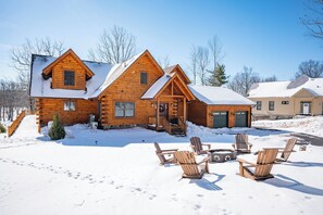 A log cabin with a snow-covered roof, a front porch, and a fire pit area with Adirondack chairs.