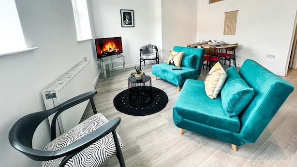A modern living room with teal sofas, a black and white chair, a fireplace, and a dining area with a table and chairs. - The County Staycation (Liverpool)