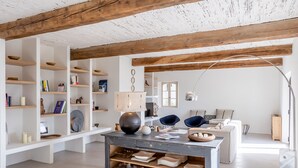 Ruime kamer met zichtbare houten balken, witte muren en een centrale tafel met kommen en boeken. De muren zijn voorzien van wandplanken, en een raam zorgt voor natuurlijk licht.