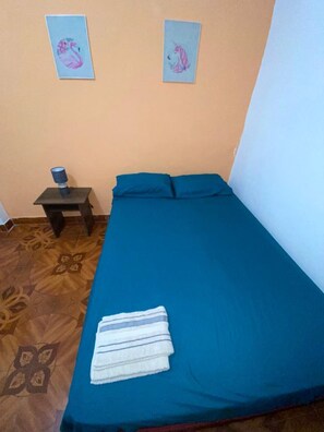 A small, neatly made bed with a blue bedspread and pillows, a small table with a lamp, and two wall-mounted paintings of swans.