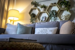 Een moderne woonkamer met een grijze bank, meerdere kussens, een lamp en honingraatplanken met planten en decoratieve objecten.