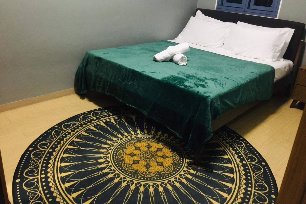 A neatly made bed with a green bedspread and white pillows, a round patterned rug, and a wooden floor.