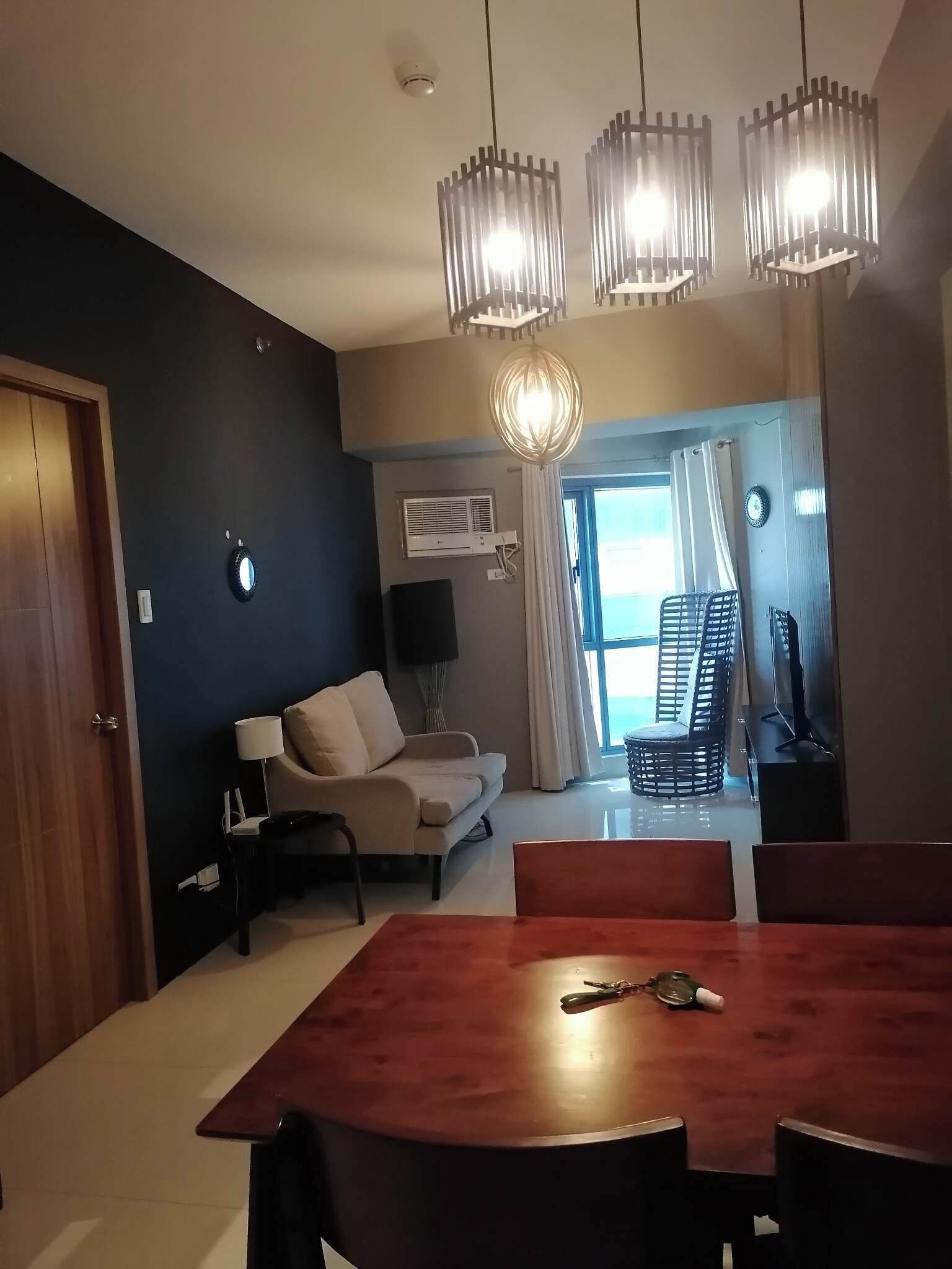A modern living room with a sofa, a dining table, and a TV. There is a window with curtains and a ceiling fan.