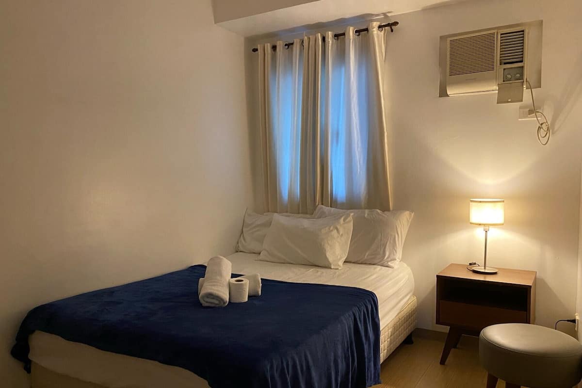 A hotel room with a bed, a nightstand with a lamp, a chair, and an air conditioning unit on the wall.