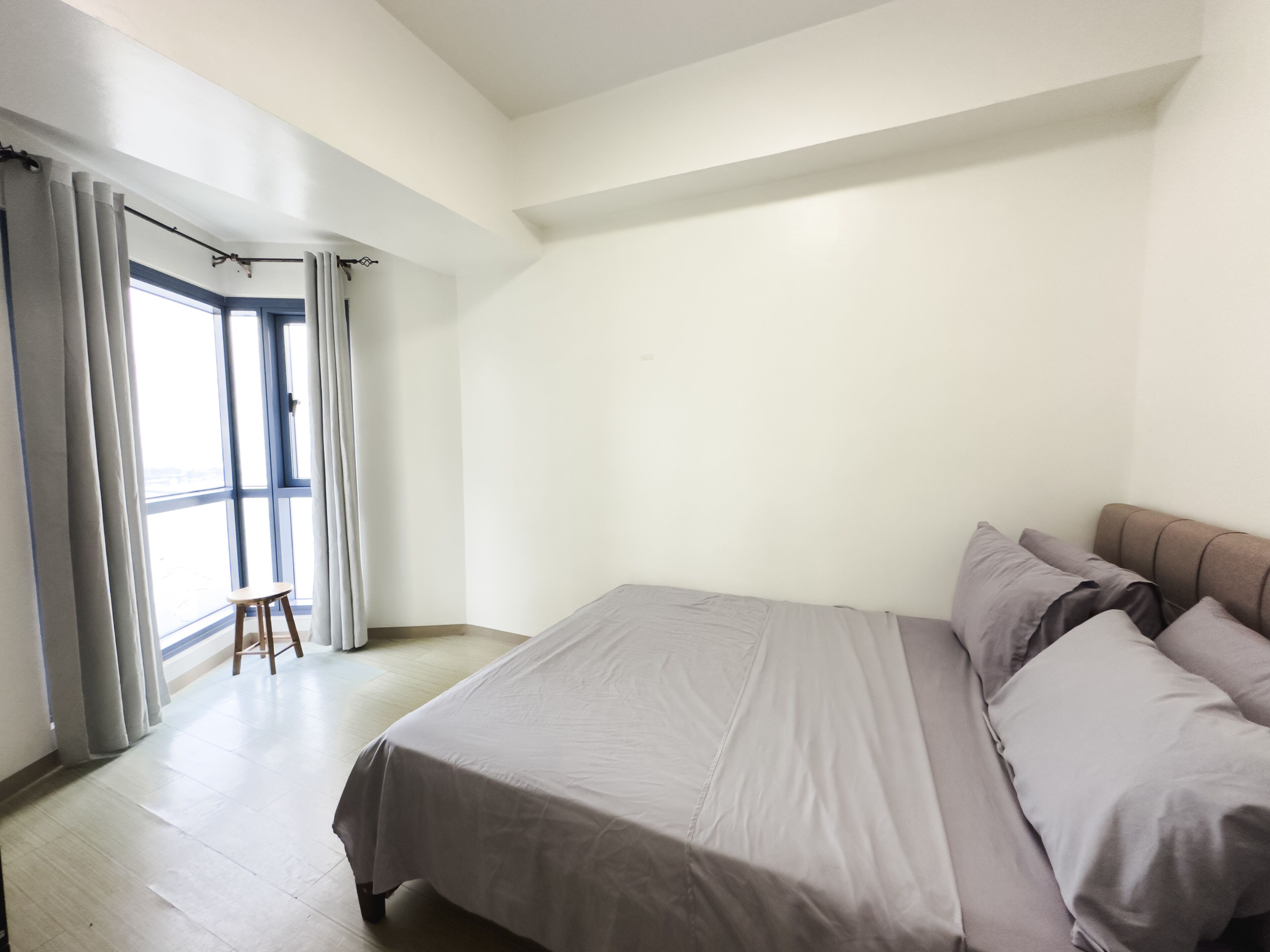 A bedroom with a large bed, a small stool, and a window with curtains.