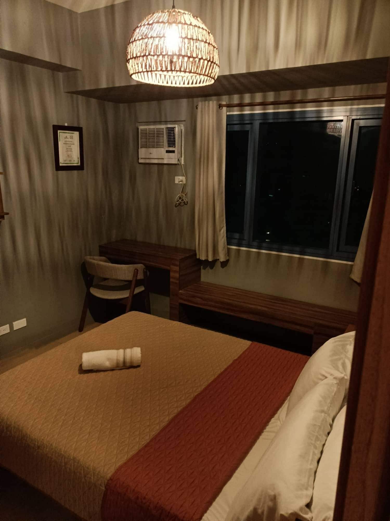 A hotel room with a bed, a desk, a chair, a window with curtains, and a hanging light fixture.