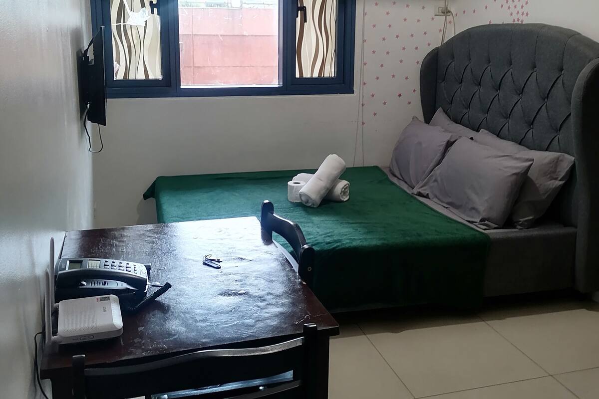 A room with a bed, a table with a phone and a speaker, a window with blinds, and an air conditioner.