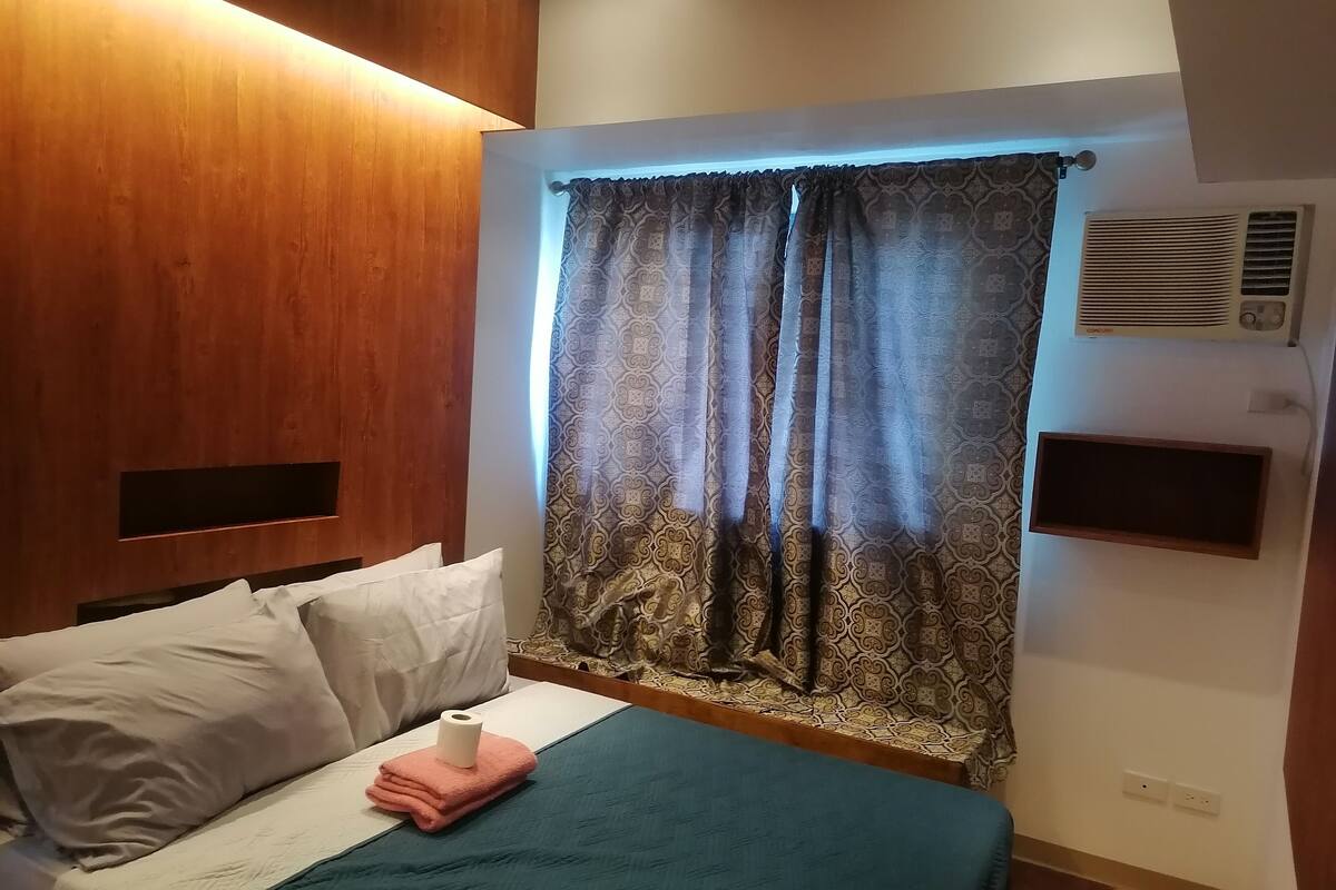 A hotel room with a bed, a wooden headboard, patterned curtains, a wall-mounted air conditioner, and a small shelf.