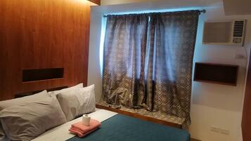 A hotel room with a bed, a wooden headboard, patterned curtains, a wall-mounted air conditioner, and a small shelf.