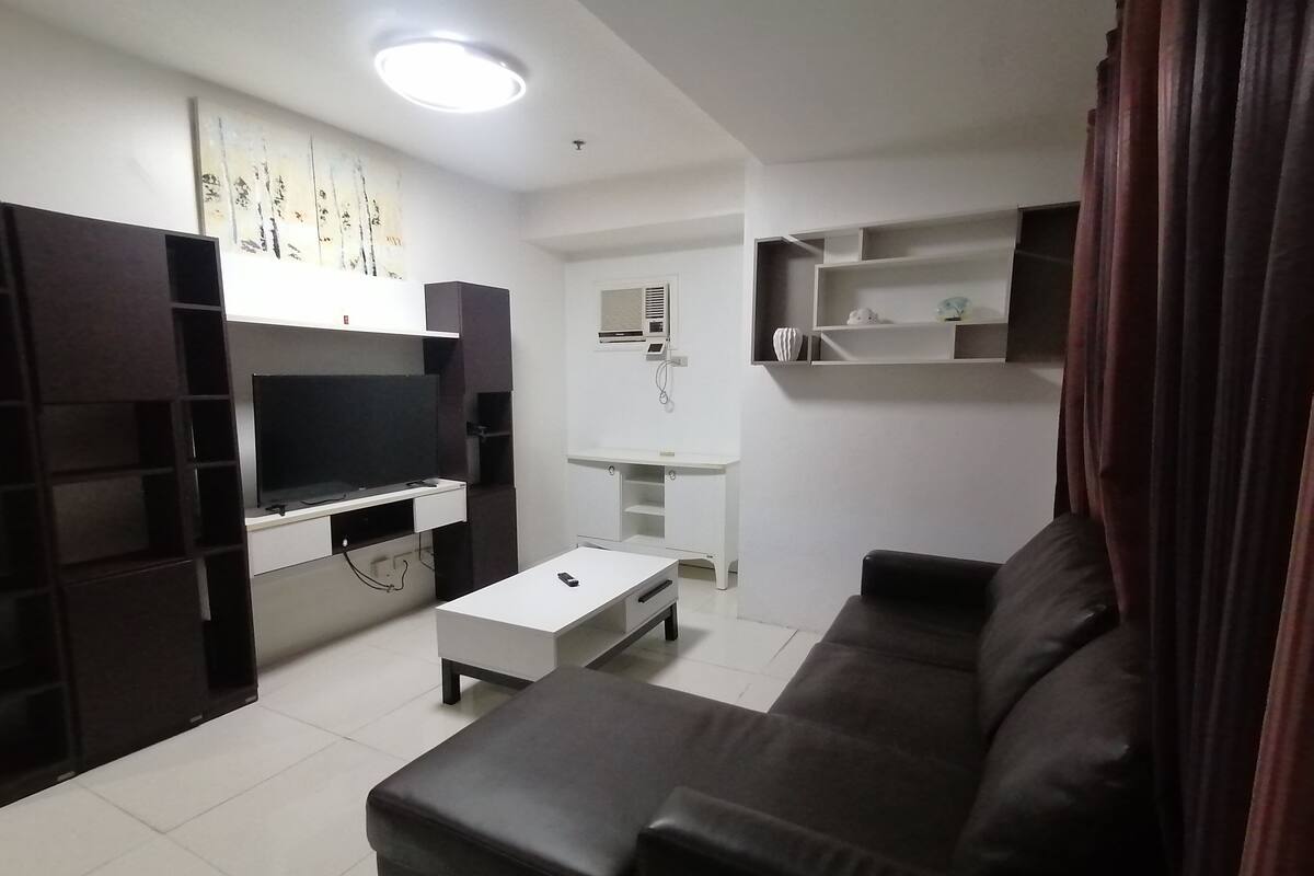 A modern living room with a flat-screen TV, a white coffee table, a black leather sofa, and a small white side table.
