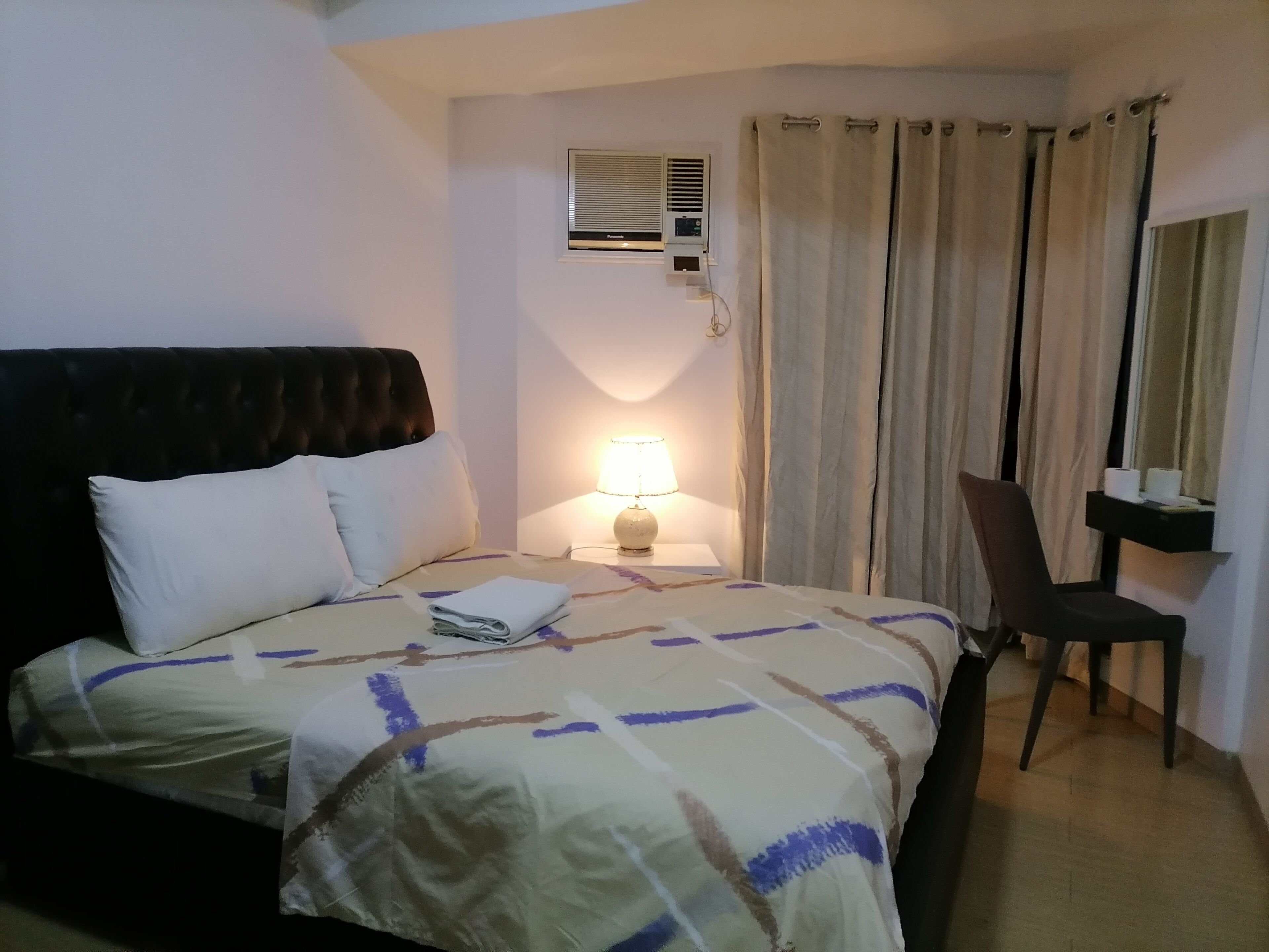 A hotel room with a bed, a desk, a chair, a lamp, and an air conditioning unit.