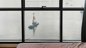 A room with a large window overlooking a calm body of water, a crane, and several boats.