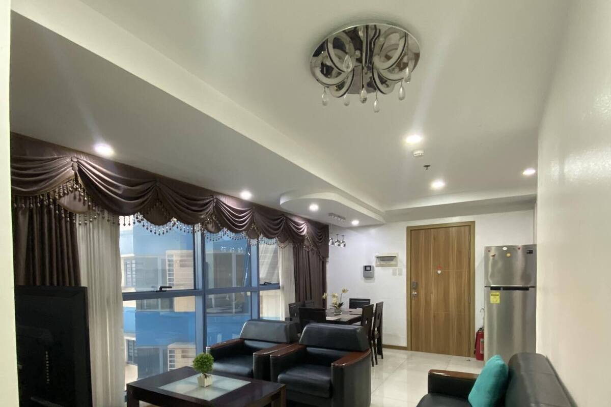 A modern living room with a sofa, coffee table, and TV. There is a dining area with chairs and a table. The room has large windows and a ceiling light fixture.