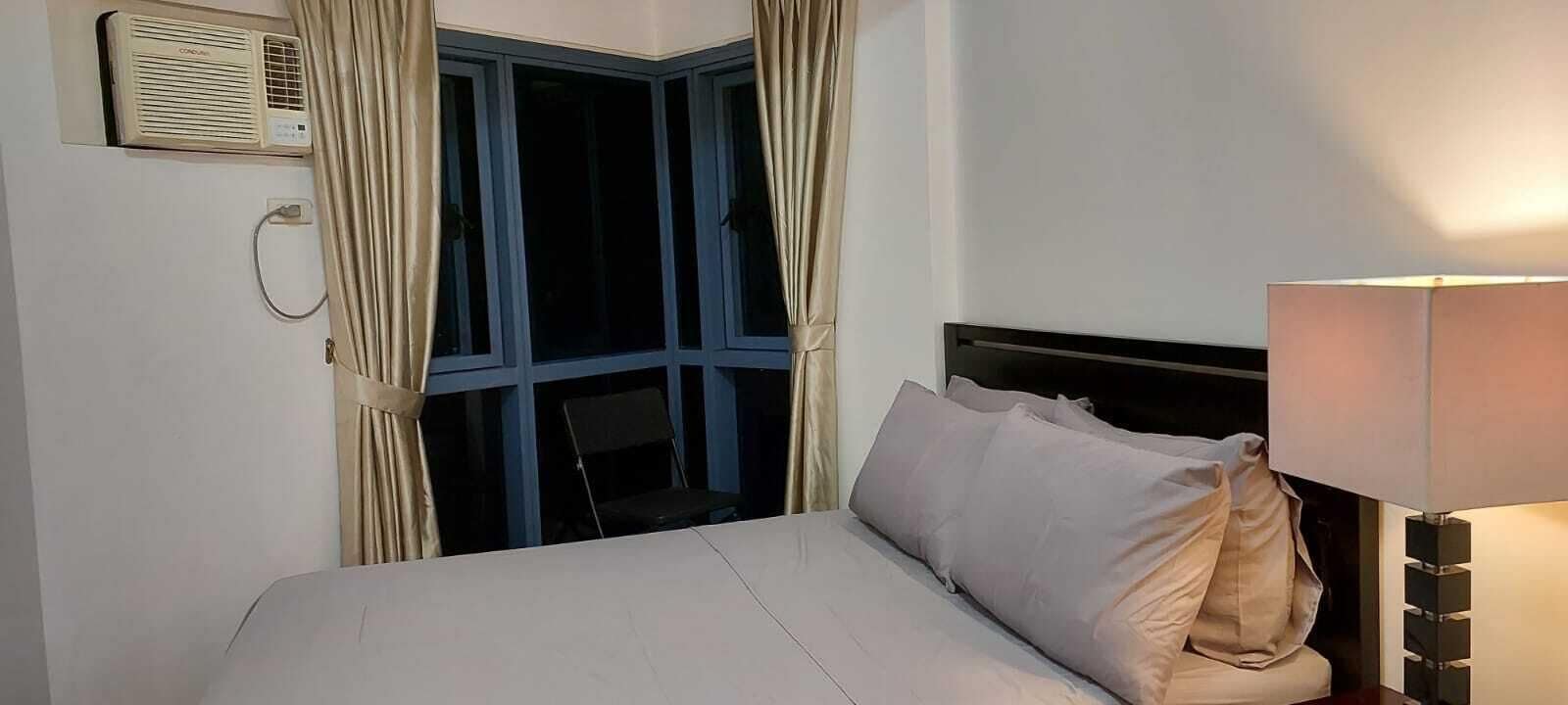 A hotel room with a bed, a window with curtains, a bedside lamp, and an air conditioning unit.