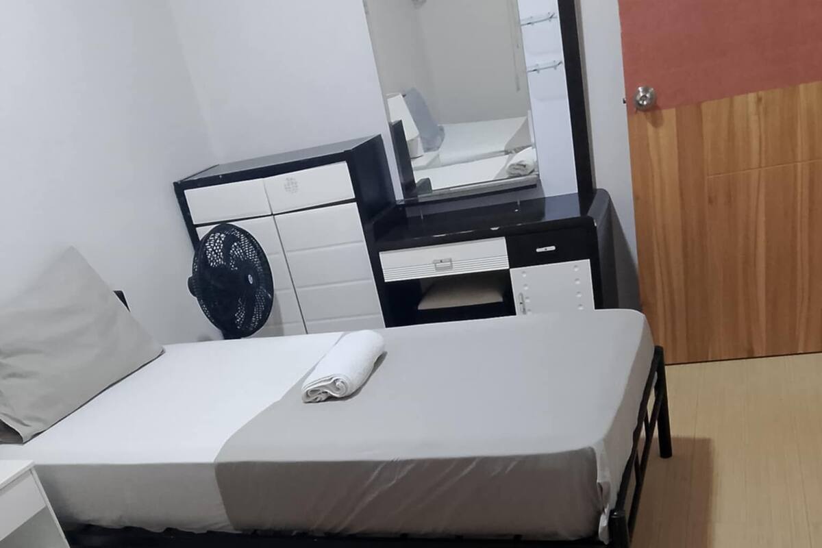 A room with two single beds, a wooden wardrobe, a desk with a mirror, and a fan.