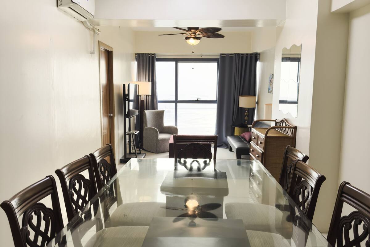 A dining area with a glass table and wooden chairs, a ceiling fan, and a view through a large window.